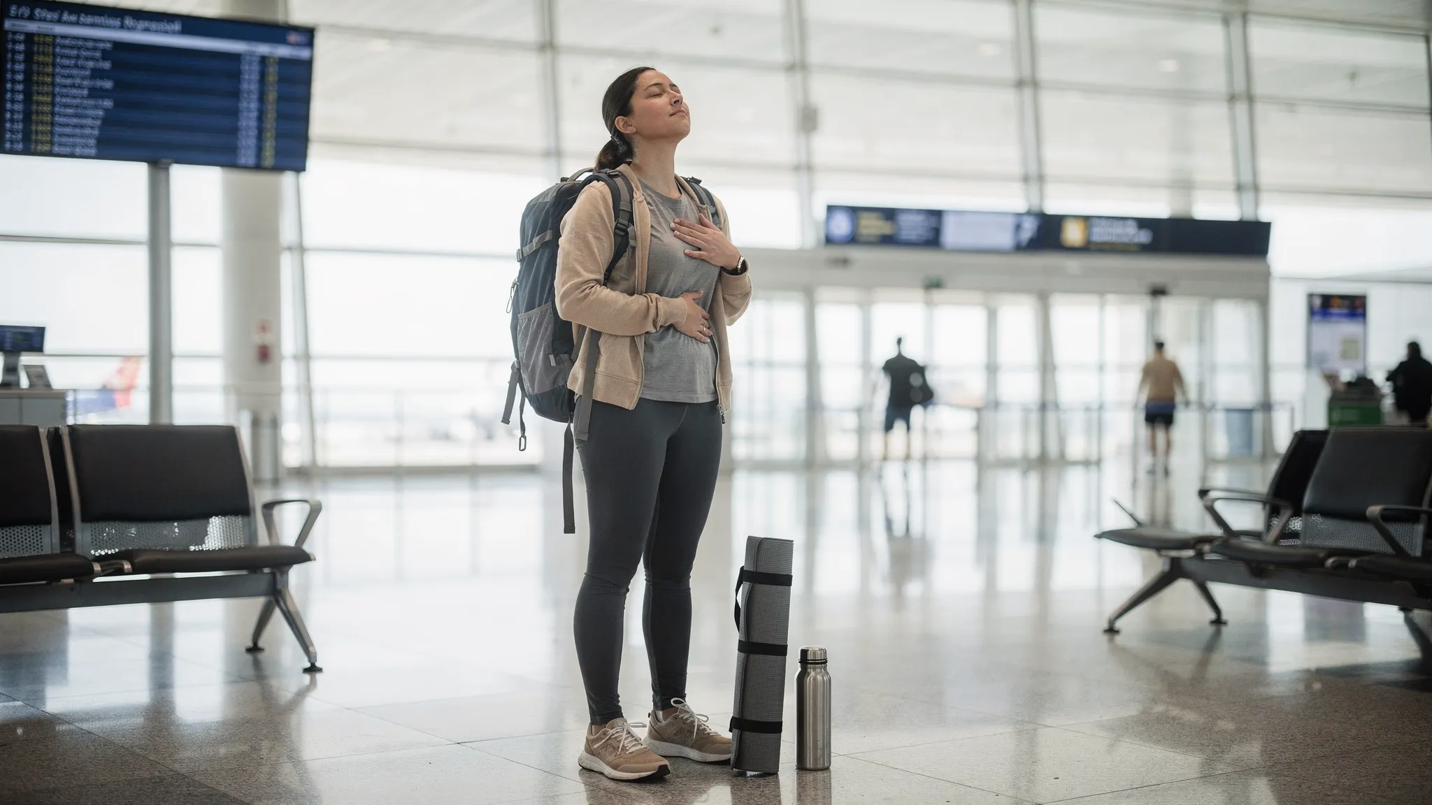 Viajera estirando y practicando respiración consciente cerca de la puerta de embarque, con mochila ligera, esterilla de viaje fina enrollada, botella reutilizable y luz natural entrando por grandes ventanales del aeropuerto.