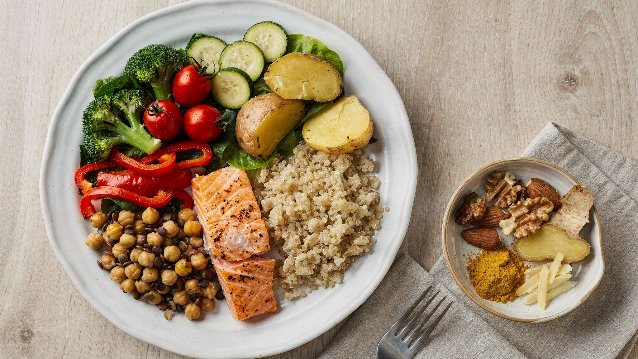 Plato mediterráneo antiinflamatorio con medio plato de verduras variadas, un cuarto de proteína (salmón o legumbres), un cuarto de carbohidrato (quinoa o patata), y un cuenco pequeño con aceite de oliva virgen extra, frutos secos y especias como cúrcuma y jengibre.