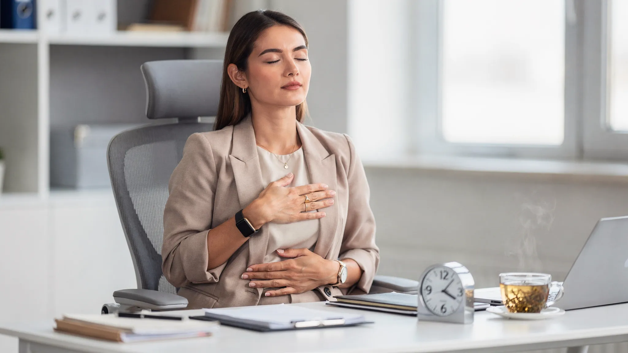 Una profesional sentada en una silla de oficina, ojos cerrados, mano sobre el abdomen y la otra sobre el pecho, postura relajada, con un reloj discreto en la mesa marcando 5 minutos y una taza de infusión al lado.