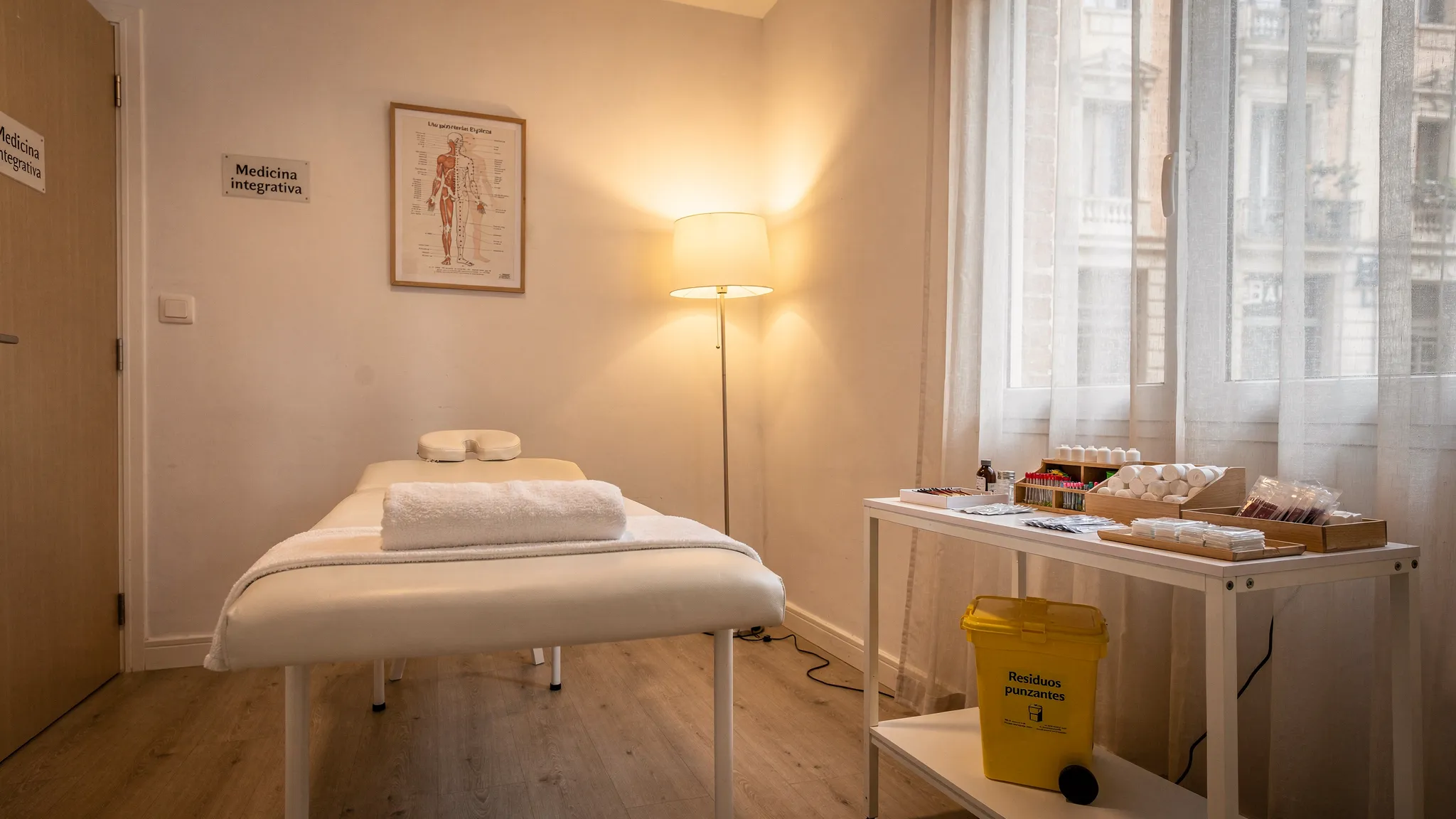 Una sala de acupuntura limpia y tranquila en Barcelona, con camilla, luz cálida, mesa auxiliar con material organizado, contenedor rígido para objetos punzantes y ambiente minimalista de consulta de medicina integrativa.