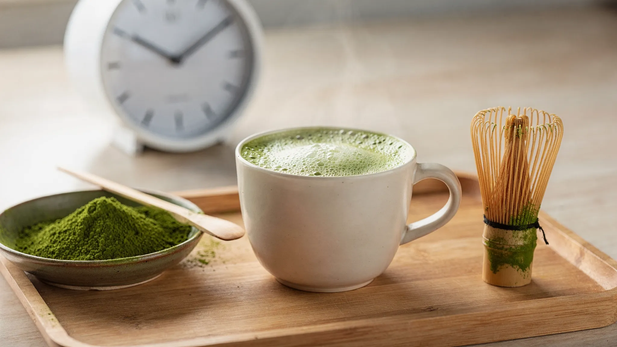 Una taza de matcha recién batido con espumado fino junto a un cuenco con polvo de matcha verde, un batidor tradicional y un reloj indicando una rutina de mañana enfocada.