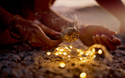 Hands, light and sage smoke at beach for healing or aromatherapy zen meditation. Woman, wellness and mental health in nature for mindfulness or spiritual chakra energy with fairy lights on ocean sand