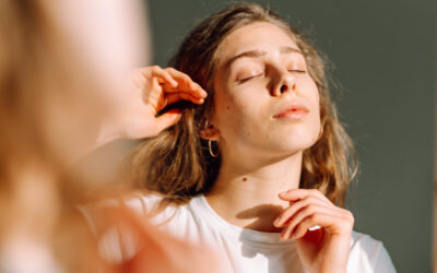 A close-up of a young woman with acne on her face looks into a mirror in a sunny room.